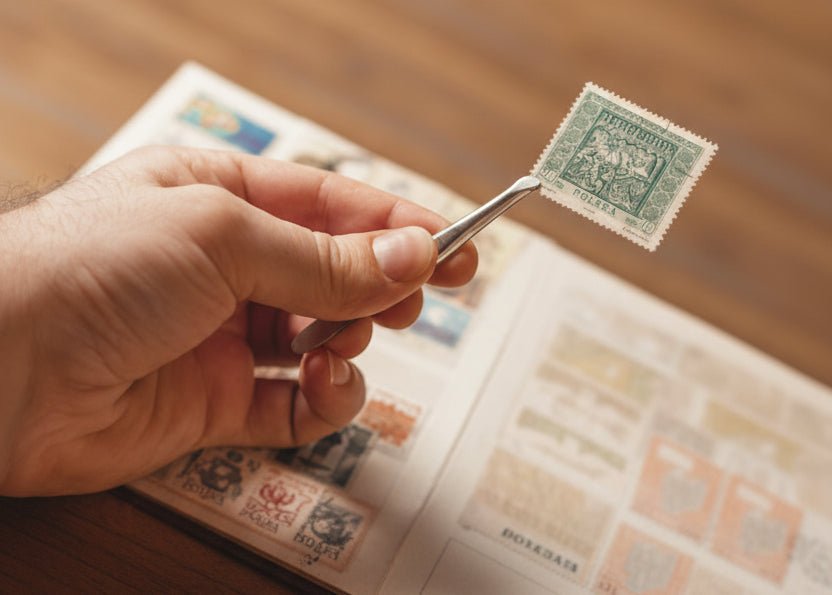 A close-up of a hand holding a single vintage blue postage stamp with tweezers over an open book of stamps on a wooden table.