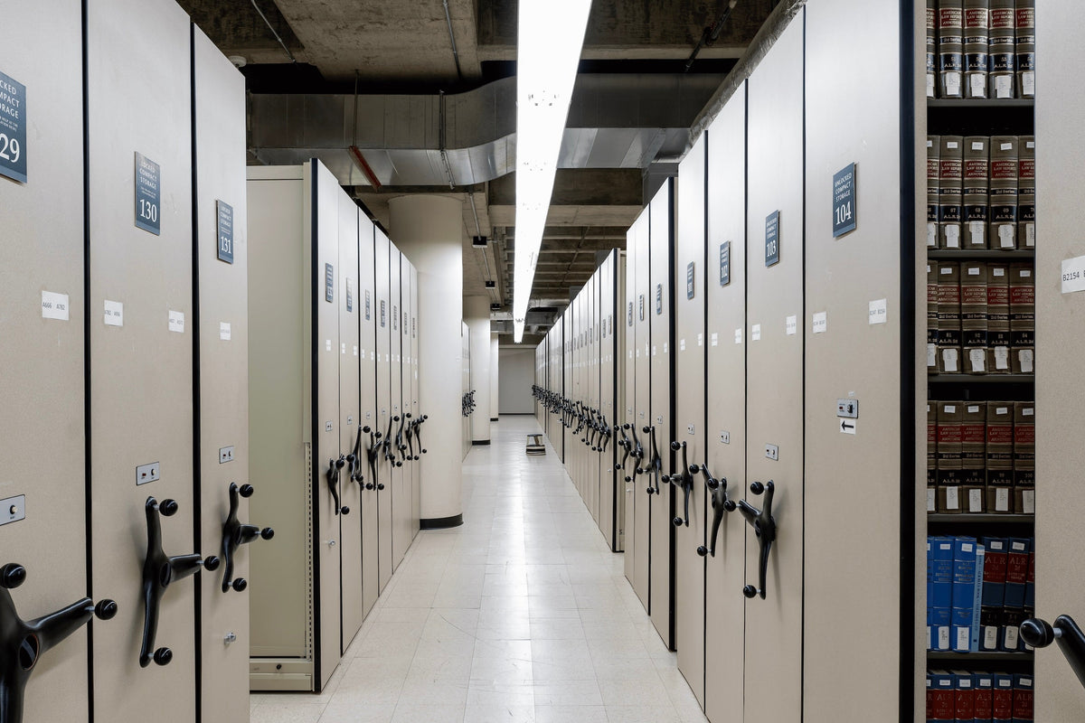 A long aisle between tall beige moving file cabinets in a library or archive, with numbered doors and a row of bound books visible on a shelf to the right.
