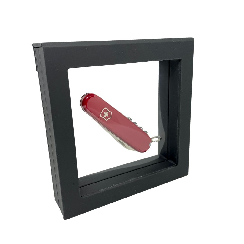 Black square frame/shadowbox with transparent window made of film that is molded to a red swiss army knife on a white background