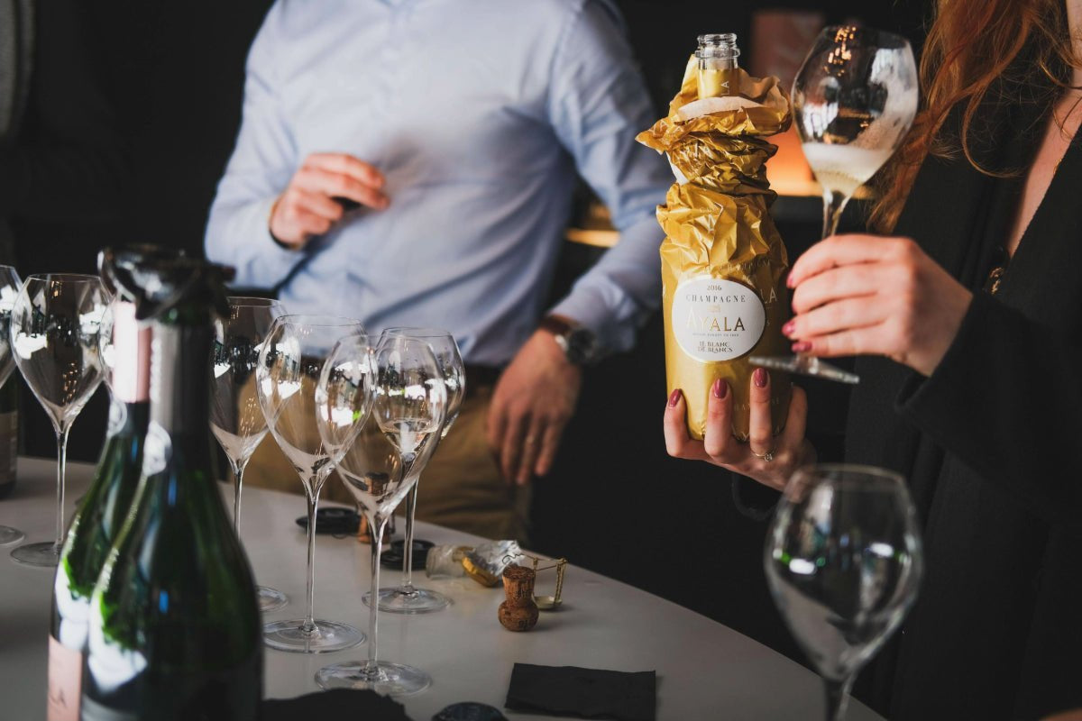 A person holds a bottle wrapped in gold foil & a wine glass. Other wine glasses and a champagne bottle sit on a table.