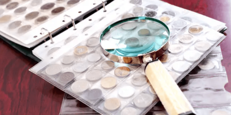 A close-up of a coin collector's binder filled with collectible coins, with a magnifying glass resting on the plastic coin trays over a dark wooden surface.