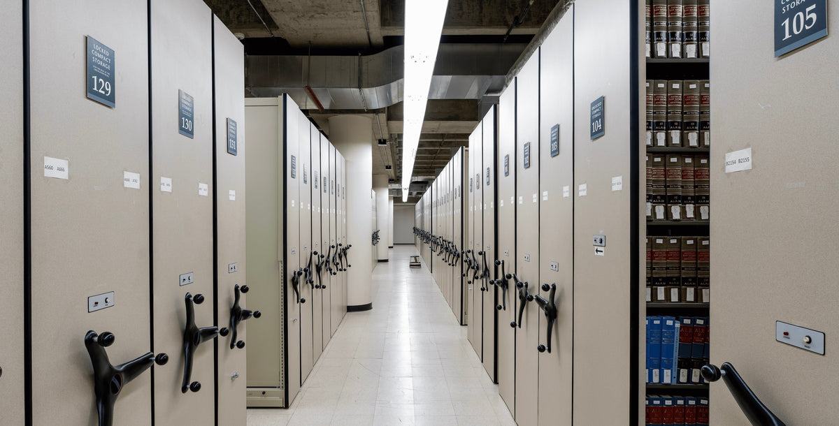 A long aisle between tall beige moving file cabinets in a library or archive, with numbered doors and a row of bound books visible on a shelf to the right.