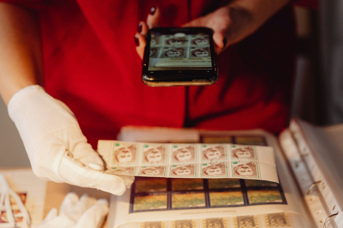 A person wearing a red shirt and white gloves holds a smartphone over a binder filled with postage stamp sheets.