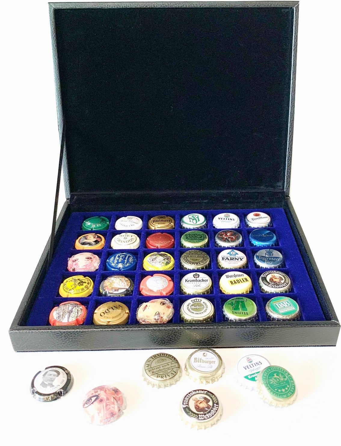 A black leather box with an open lid, lined with blue velvet and divided into compartments holding many colorful bottle caps. Several bottle caps are placed in front of the box on a white background.