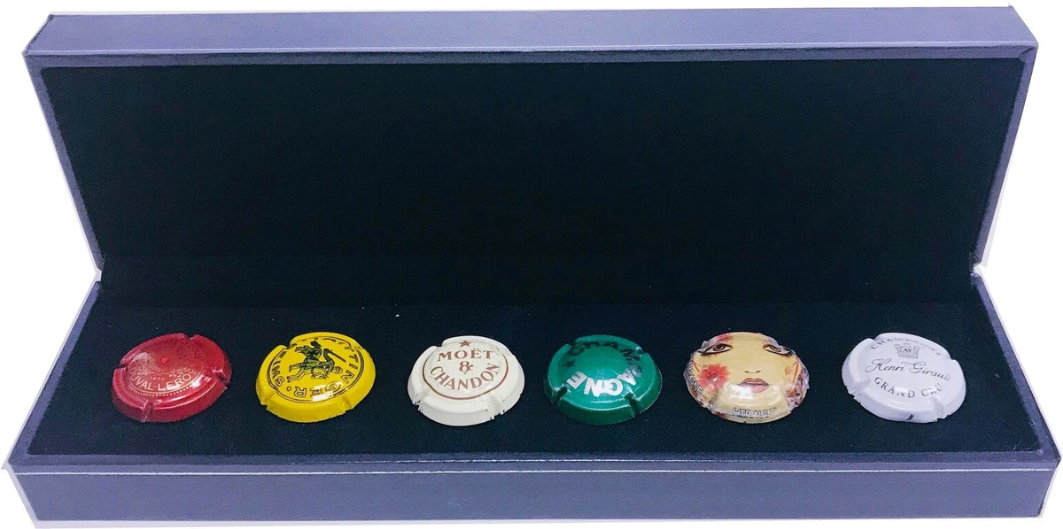 Six champagne caps displayed in a dark blue presentation box. The caps are red, yellow, white, green, beige, and white, with some featuring visible brand text such as 'Moget de Champagne' and 'Mons'.