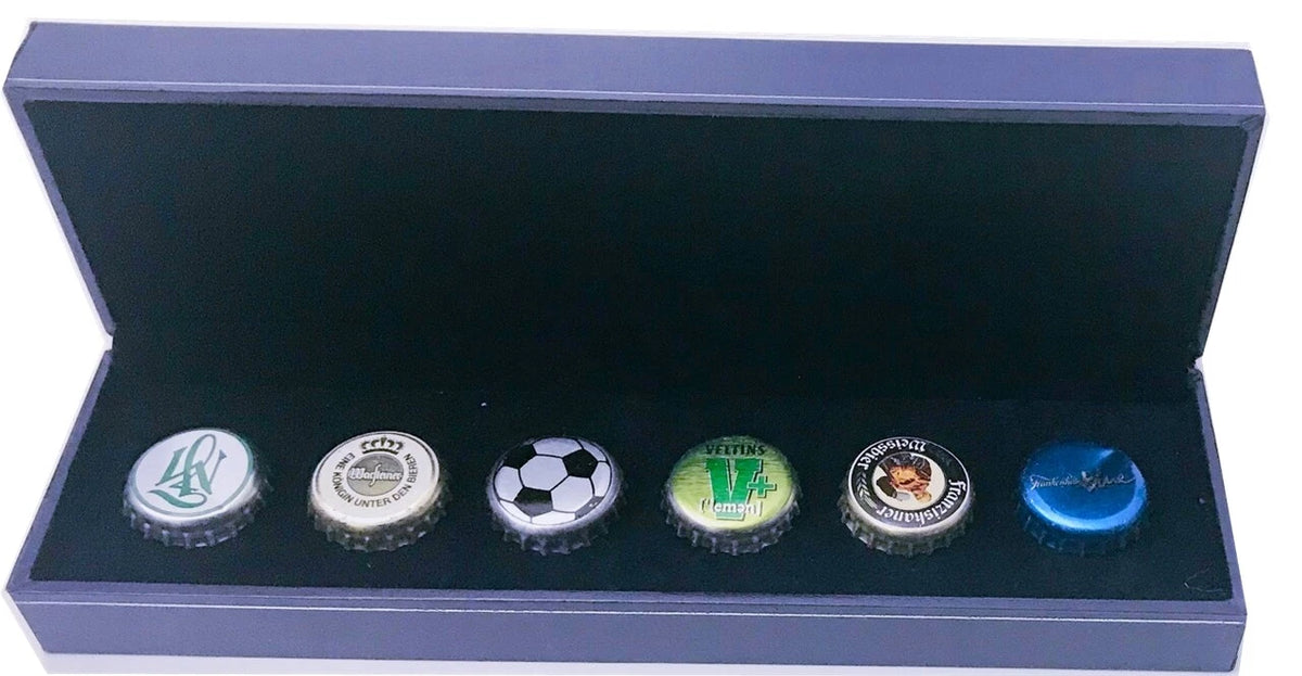 A set of six small bottle caps displayed inside an open dark presentation box. The bottle caps feature various designs, including sports team logos, a soccer ball pattern, and a green 'W' emblem.