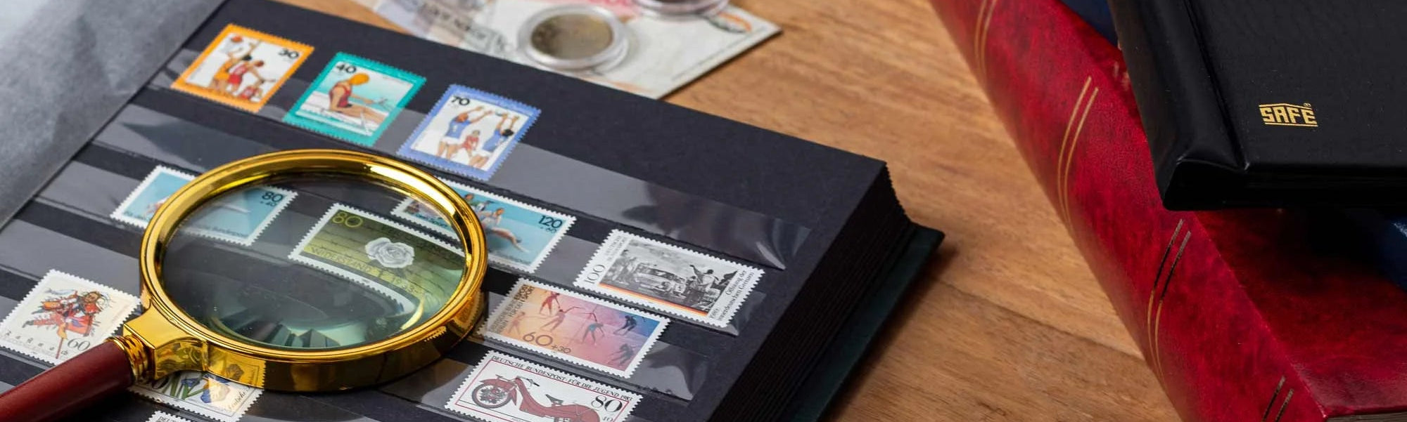 A close-up of a black postage stamp album on a wooden desk, with a gold-rimmed magnifying glass resting on the album cover. Several stamps are visible inside the album, with additional stamps, banknotes, and stacked books nearby.