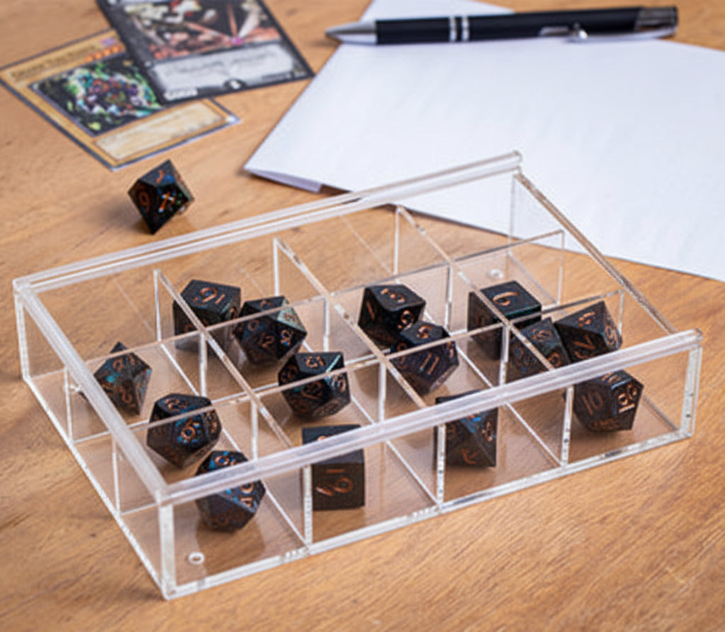 A clear acrylic organizer tray with multiple compartments holding several black polyhedral gaming dice, placed on a wooden tabletop with cards, a pen, and papers in the background.